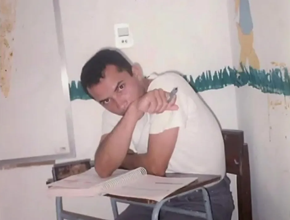 Foto antiga de uma sala de aula, com um jovem apoiado na carteira e um caderno aberto
