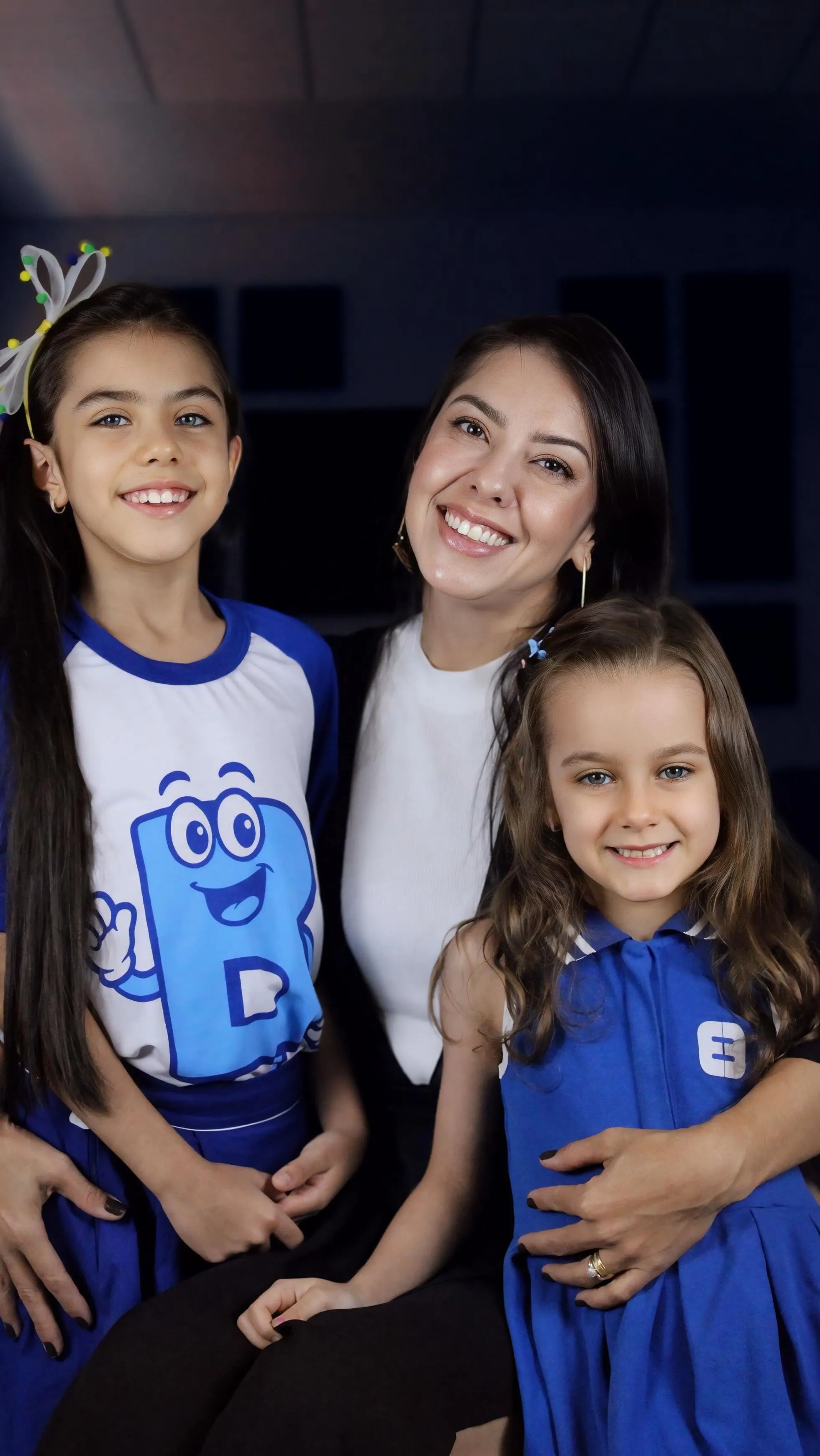 Mãe abraçada às duas filhas de uniforme, ao lado do B, o mascote azul do Educandário Bonus