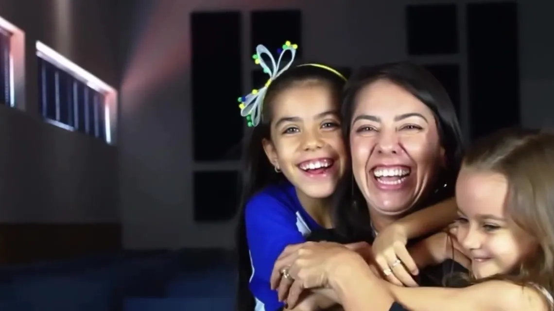Mãe sorrindo e abraçando as duas filhas durante a gravação do depoimento