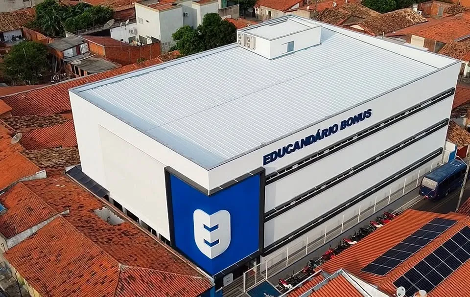 Vista aérea da sede do Educandário Bonus: prédio branco de três andares com a torre azul da marca, cercado pelos telhados de Água Branca